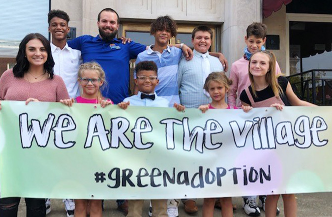 The Green family all together and holding a banner that says, "We are the village."